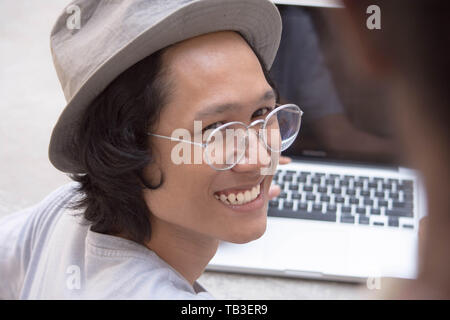 Giovane uomo asiatico con cappello e occhiali sorridendo a un amico con il computer portatile in background Foto Stock