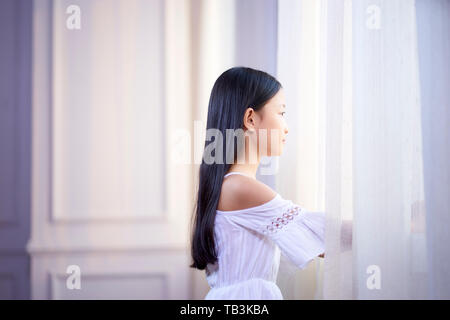 Bella ragazza asiatica con lunghi capelli neri guardando fuori della finestra in camera da letto Foto Stock