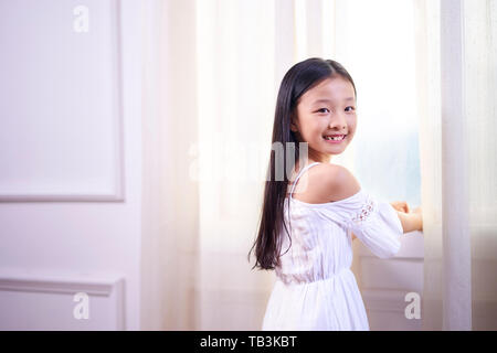 Bella ragazza asiatica con lunghi capelli neri in piedi di fronte a windwo passando per la fotocamera a sorridere Foto Stock