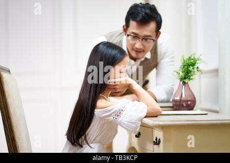 Triste ragazza asiatica seduto alla scrivania nella sua stanza e ottenere il comfort da padre amorevole Foto Stock