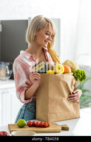 Felice donna bionda holding credito carta vicino al sacchetto di carta con negozi di generi alimentari Foto Stock