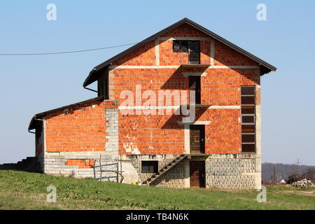 Incompiuta di grandi dimensioni in mattoni rossi suburban casa famiglia sul lato della piccola collina circondato da erba non tagliata e cielo blu chiaro in background Foto Stock
