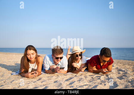 Gruppo di millenials smartphone utilizzando la posa insieme sul telo da spiaggia vicino al mare in estate il tramonto. I giovani tossicodipendenti da mobile smart phone. Sempre Foto Stock