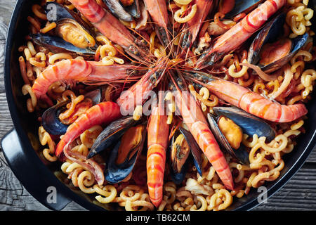 Fideua, una paella come piatto con pasta speciale, gamberoni, bianco carne di pesce, calamari, cozze in una padella nera su una tavola di legno, vista da sopra, fla Foto Stock