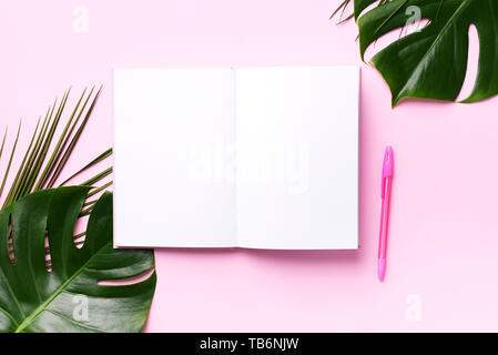 Green monstera, foglie di palma, di cocco, di notebook su sfondo rosa. Femmina scrittore blog workspace, torna al concetto di scuola. Lay piatto. Vista superiore Foto Stock