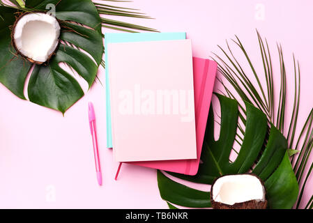 Green monstera, foglie di palma, di cocco, di notebook su sfondo rosa. Femmina scrittore blog concetto di area di lavoro. Lay piatto. Vista superiore Foto Stock