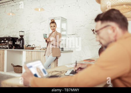 Pausa caffè. Bella Donna che mantiene il suo smartphone e una tazza di caffè in piedi vicino alla macchina del caffè e guardando il suo boss. Foto Stock