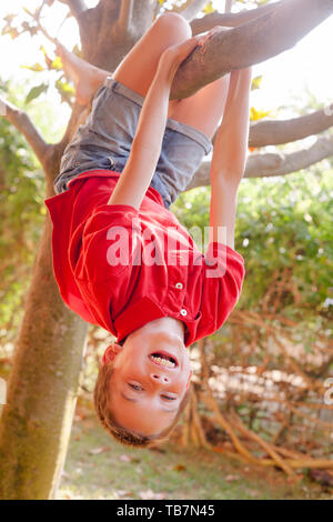 Ritratto di ragazzo spensierato appeso a testa in giù da un albero in un parco godendo di estate Foto Stock