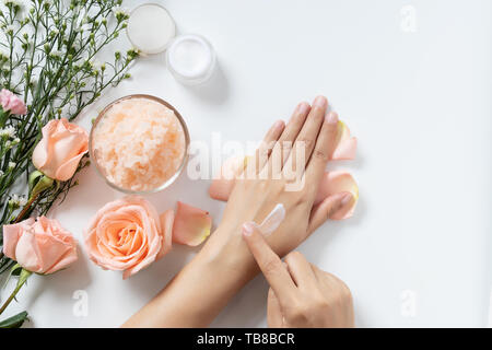 Dermocosmesi naturale concetto. donna applicare la crema bianca su le mani su sfondo bianco con un vasetto di crema cosmetica, sale spa scrub ,rose e fiori di colore bianco Foto Stock