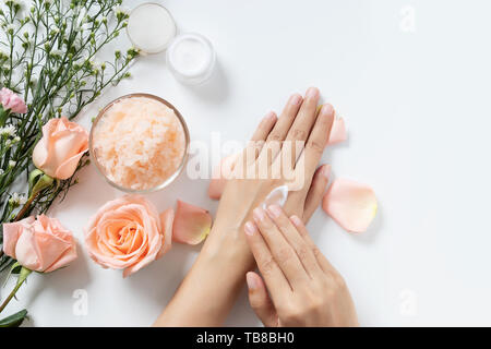 Dermocosmesi naturale concetto. donna applicare la crema bianca su le mani su sfondo bianco con un vasetto di crema cosmetica, sale spa scrub ,rose e fiori di colore bianco Foto Stock