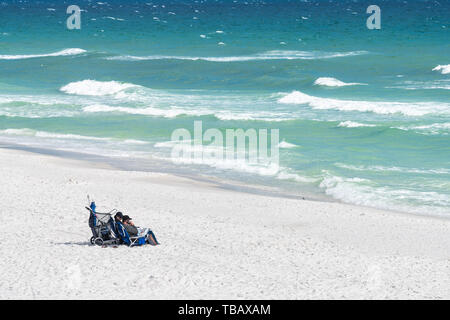 Destin, Stati Uniti d'America - 24 Aprile 2018: Miramar Beach al Golfo del Messico con coppia seduti su sedie a prendere il sole sulla sabbia di mare oceano onde si infrangono in estate Foto Stock