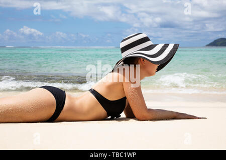 Vista laterale di una giovane donna in Bikini indossando Striped Hat giacente sulla spiaggia Foto Stock