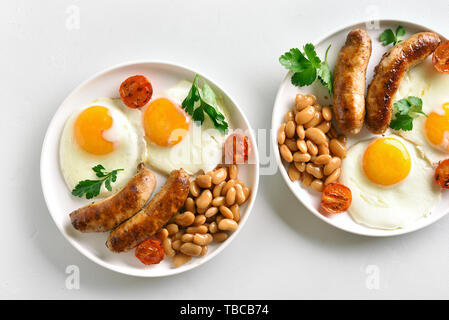 La prima colazione con uova fritte, salsicce, fagioli, pomodori verdi su piastra sulla pietra bianca dello sfondo. Vista superiore, laici piatta Foto Stock