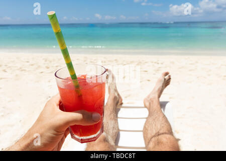 Close-up di un uomo disteso sulla spiaggia tenendo un bicchiere di succo rosso in mano Foto Stock