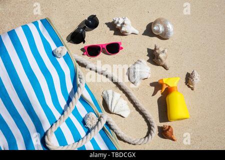 Conchiglie, occhiali da sole e crema solare, bottiglia e borsa sulla sabbia in spiaggia Foto Stock