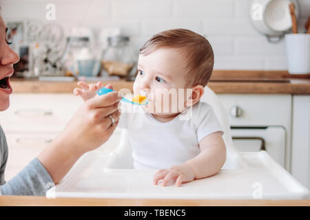 Affascinante piccolo bimbo 6-8 mesi prima di mangiare cibo da zucca cucchiaio con la mamma a casa Foto Stock