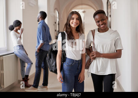Piuttosto africano e gli studenti caucasici ragazze in piedi sul corridoio di università e sorridente alla fotocamera. Le donne mantenendo gli amici, studiare in università internazionale insieme. Foto Stock