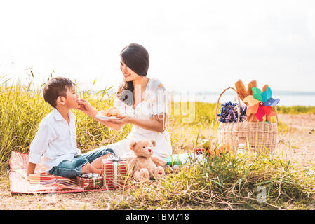 Bella asiatica mom snack di alimentazione a suo figlio in prato quando fare picnic. Madre e figlio giocare insieme. Celebrando nella Madre di giorno e di apprezzare Foto Stock