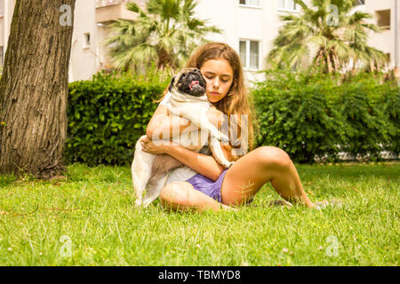 Giovane ragazza adolescente avvolge il suo cane pug nel parco su erba verde Foto Stock