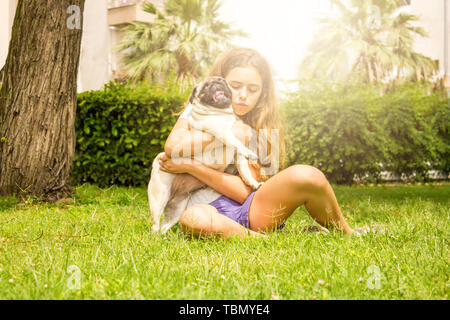 Giovane ragazza adolescente avvolge il suo cane pug nel parco su erba verde Foto Stock