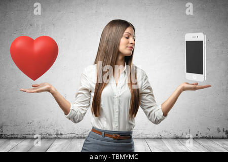 Young business woman holding cuore rosso e smartphone nelle sue mani sul muro grigio sfondo Foto Stock