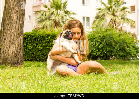 Giovane ragazza adolescente avvolge il suo cane pug nel parco su erba verde Foto Stock