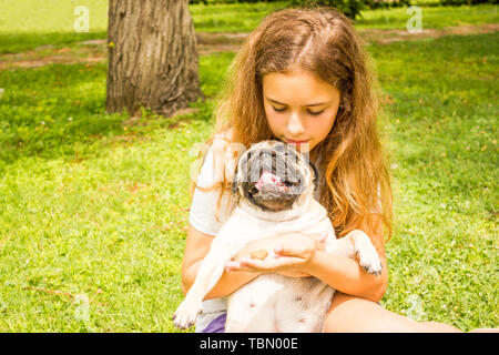 Giovane ragazza adolescente avvolge il suo cane pug nel parco su erba verde Foto Stock