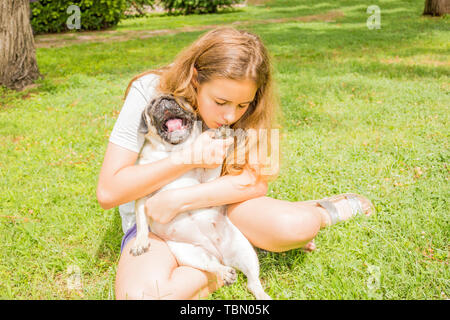 Giovane ragazza adolescente avvolge il suo cane pug nel parco su erba verde Foto Stock