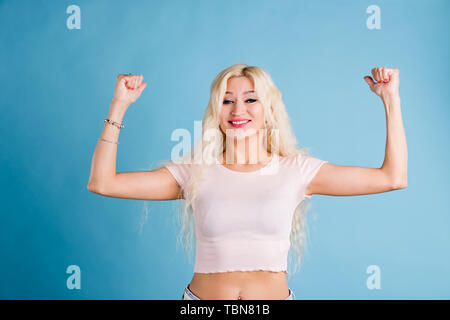 Io sono felice vincitore fiducioso bella donna con capelli biondi, alzando le mani e mostra i muscoli, dimostrando la sua forza, indossando tee shirt isolato o Foto Stock