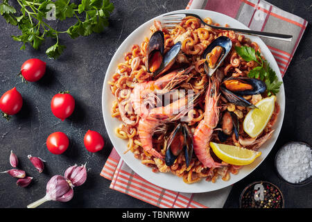 Fideua spagnola, un noodle Paella con gamberoni, bianco carne di pesce, calamari, cozze servite su una piastra bianca con una forcella di argento, su un cemento grigio ta Foto Stock