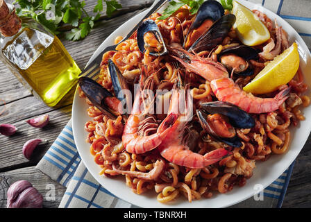 Close-up di Fideua spagnola, un noodle Paella con gamberoni, bianco carne di pesce, calamari, cozze servite su un piatto bianco su un tavolo di legno con cartine Foto Stock