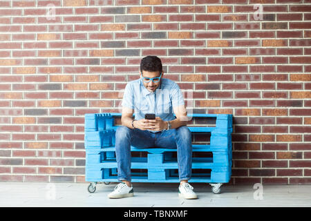 A piena lunghezza Ritratto di successo del bel giovane uomo barbuto in stile casual, occhiali seduto su blu pallet di legno, azienda, guardando al mobile sm Foto Stock