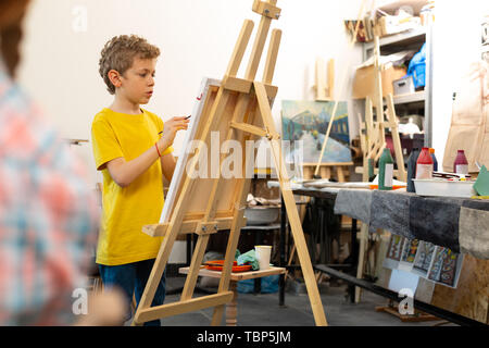 In giallo t-shirt. Biondo e talentuoso ragazzo indossa giallo t-shirt dipinto sul cavalletto di disegno Foto Stock