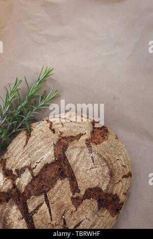 Bio grano intero pane casereccio realizzato sulla pasta acida posa su carta kraft vicino al rosmarino, girato in stile rustico. Foto Stock