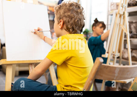 Scolaro disegno. Curly biondo e schoolboy giallo che indossa la t-shirt disegno nella scuola di arte Foto Stock