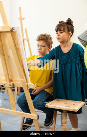 Ragazza carina aiutando. Carino ragazza dai capelli scuri aiutando la sua bionda compagna di scuola di pittura alla scuola d'arte Foto Stock