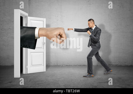 Grande pugno teso che appare attraverso la porta bianca e imprenditore, rendendo la calciata gesto sul muro grigio sfondo Foto Stock