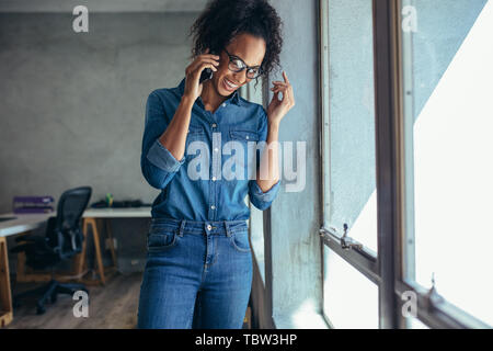 Imprenditrice parlando al telefono cellulare e sorridente. Imprenditore femmina in piedi da una finestra e parlando su telefono cellulare in ufficio Foto Stock