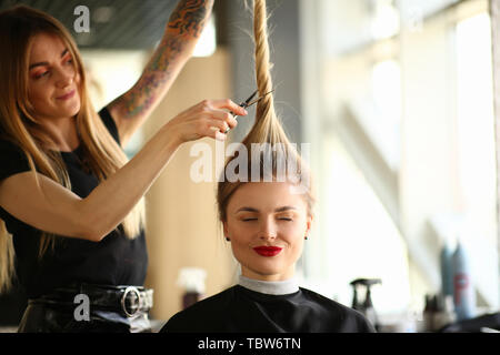 Tatuato Stylist il taglio di capelli di occhi chiusi donna. Parrucchiere con le forbici per fare l'acconciatura per Client femmina. Ragazza bionda Getting Haircut in Foto Stock