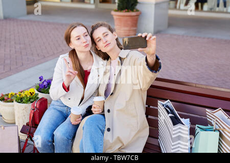 Elegante del contenuto delle giovani donne in abiti casual seduta sul banco di lavoro con tazzine di caffè e posa per selfie realizzata sullo smartphone Foto Stock