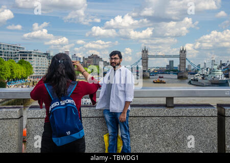 Una signora prende una fotografia del suo compagno sul Ponte di Londra con il fiume Tamigi, HMS Belfast e Tower Bridge in background. Foto Stock