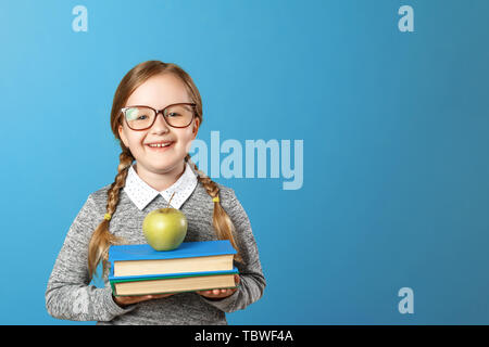 Ritratto di un allegro bambina con gli occhiali su uno sfondo blu. La scolaretta è in possesso di una pila di libri e un apple. Si torna a scuola. Il concetto Foto Stock