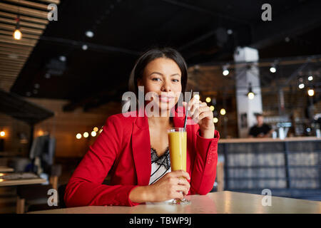 Ritratto di contenuto giovane e bella donna nera in giacca rossa seduta a tavola nel loft bar e cocktail di agitazione con paglia Foto Stock