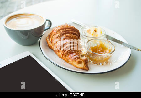 Freschi croissant croccante con marmellata, burro e cappuccino con belle latte art sulla tabella grigia in una caffetteria. Foto Stock