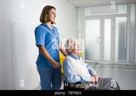 Premurosa infermiera spingendo senior paziente in sedia a rotelle all'ospedale Foto Stock