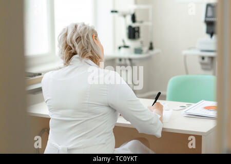 Esperto medico oculista bionda. Focalizzata a pelo corto medico in veste bianca seduta al tavolo e scrivere le raccomandazioni Foto Stock