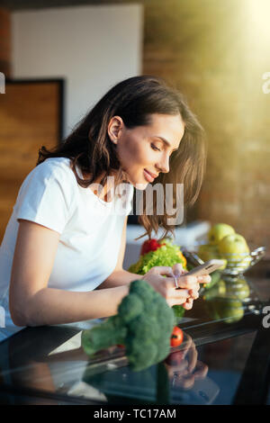 Ritratto di lei bella attraente ragazza deliziosa ricetta di lettura online su telefono cellulare fresca cena pranzo Biologico di fattoria vegs in luce interio bianco Foto Stock
