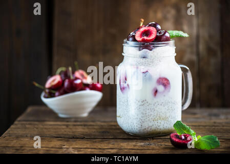 Luce a livelli sani budino dessert da Chia semi nel latte di mandorla con yogurt e ciliegie nel vasetto di vetro nel buio di sfondo di legno Foto Stock