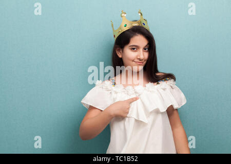 Giovani arroganza bella principessa brunette girl usura in abito bianco e diadema dorato corona, permanente e puntare il dito per se stessa, guardando è venuto Foto Stock