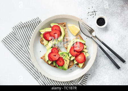 Avocado toast alla fragola su una piastra. Tabella vista dall'alto su sfondo di calcestruzzo. Vegano sano, vegetariane prima colazione o uno spuntino Foto Stock